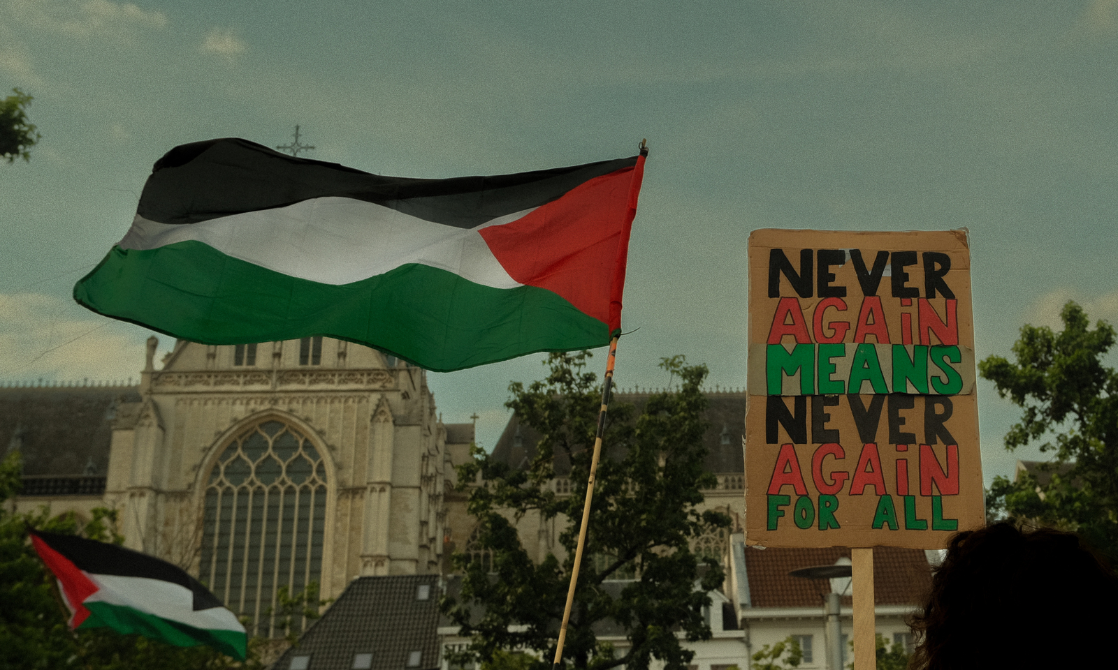 Een Palestijnse vlag wappert naast een protestbord waarop "never again means never again for all" op staat. In de achtergrond zie je de kathedraal van Antwerpen
