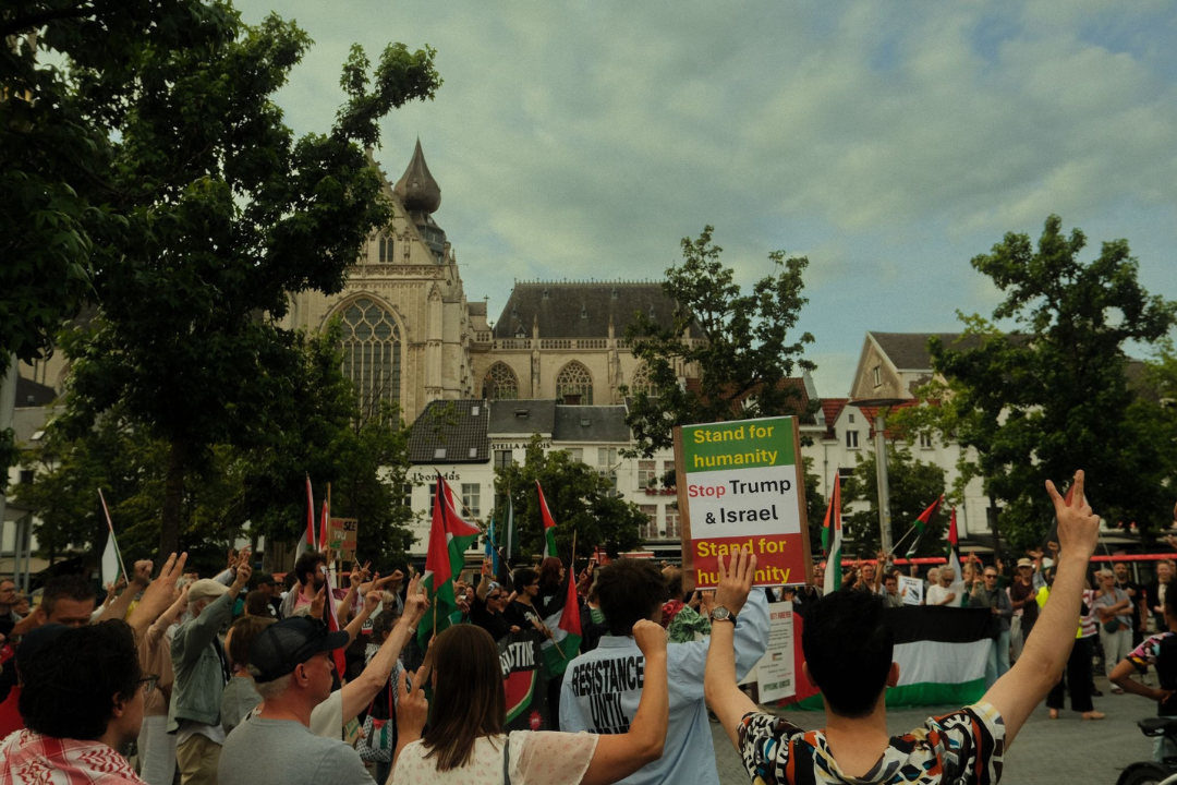 De foto toont een Antwerpse pro Palestina protest voor met de kathedraal van Antwerpen op de achtergrond. De protesters dragen Palestijnse vlaggen en er staat 1 bord op de afbeelding waarop hetvolgende staat 'Stand for humanity, stop Trump & Israel, Stand for humanity'
