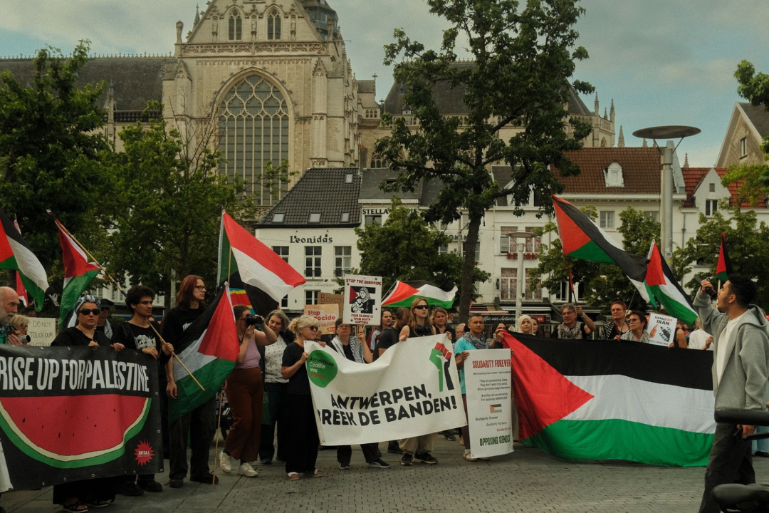 Eeen foto van pro Palestinaprotesten met dee kathedraal van Antwerpen op de achtergrond. Naast veel Palestijnse vlaggen houden mensen ook twee grote banners met tekst op vast. Op de linkse banner staat "Rise up for Palestine', met de afbeelding van een watermeloen op de achtergrond. Op de middelste banner staat 'ANTWERPEN, BREEK DE BANDEN!'. De rechtse banner is een Palestijnse vlag. Voor de deelnemers staat ook een jonge man in een microfoon protestleuzen te roepen.